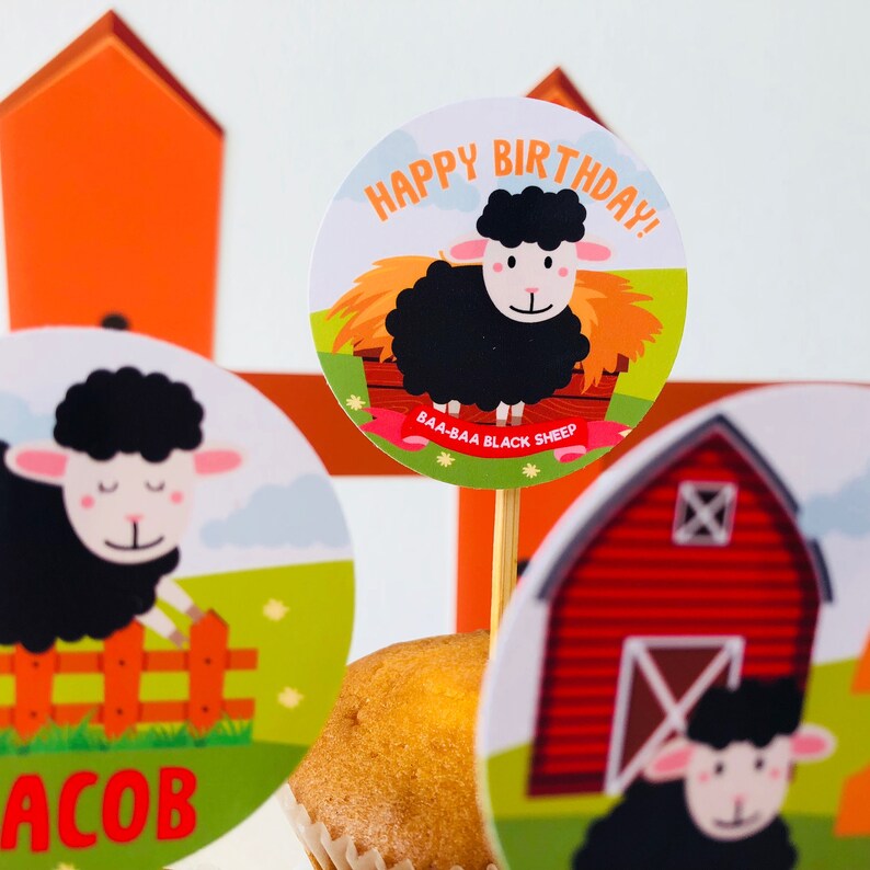 Puede incluir: Un conjunto de adornos para cupcakes con una oveja negra con el texto "Happy Birthday!" y "Baa-Baa Black Sheep" en una pancarta roja y blanca. Los adornos est&aacute;n sobre un cupcake con glaseado marr&oacute;n.