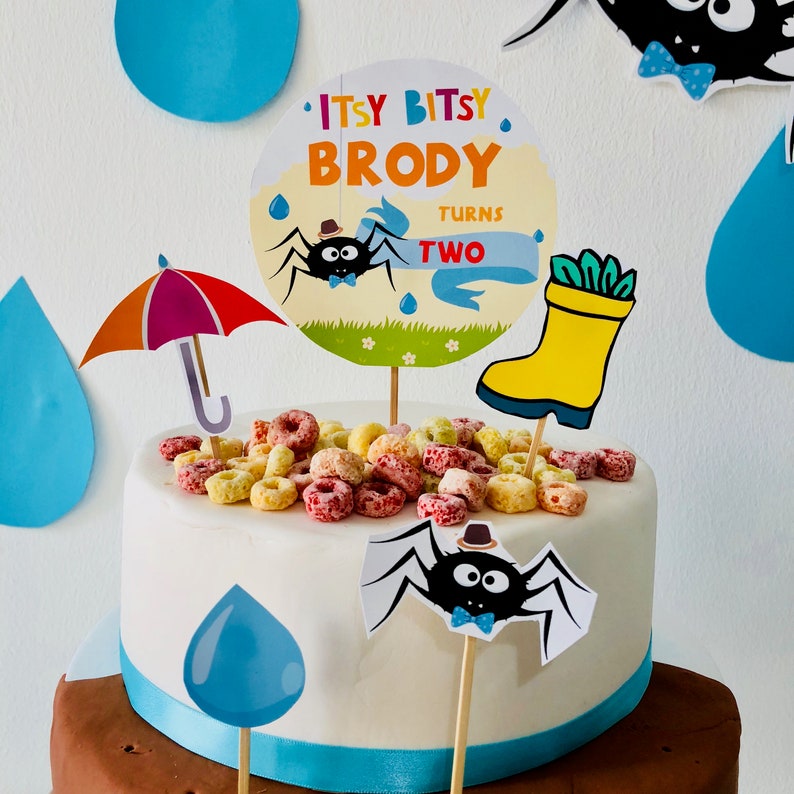 May include: A white cake decorated with colorful cereal, a blue and white rain drop, and a spider with a top hat. The cake is topped with a sign that says "Itsy Bitsy Brody Turns Two".