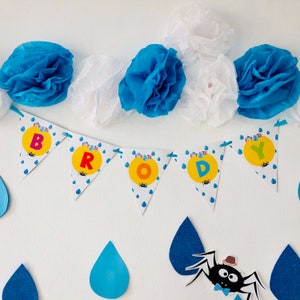 May include: A blue and yellow banner with the word "BRODY" spelled out in white letters. The banner is decorated with raindrops and a spider. The banner is hanging from blue and white tissue paper pom-poms.
