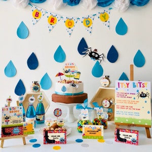 May include: A birthday party table setting with a blue and green color scheme. The table features a cake with a spider and umbrella topper, a sign with the words "Itsy Bitsy" and a spider, and various other decorations, including candy, drinks, and food. The table is decorated with blue and green raindrops and a banner that says "Brody".