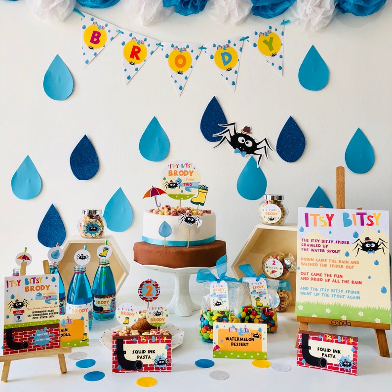 May include: A birthday party table setting with a blue and green color scheme. The table features a cake with a spider and umbrella topper, a sign with the words "Itsy Bitsy" and a spider, and various other decorations, including candy, drinks, and food. The table is decorated with blue and green raindrops and a banner that says "Brody".