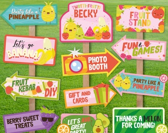 May include: A collection of colorful party signs on wooden stakes. The signs feature fruit-themed illustrations and phrases like "Party like a Pineapple," "Fruit Stand," and "Photo Booth." The signs are set against a green grassy background.