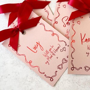 May include: Three pink cards with red ribbon bows and the text "Lucy Will you be my Maid of Honor?"  "Kamilla Will you be my Bridesmaid?" and "[unclear text]"