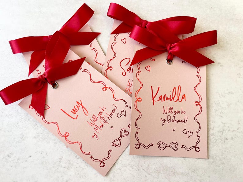 May include: Three pink cards with red ribbon bows and the text "Lucy Will you be my Maid of Honor?"  "Kamilla Will you be my Bridesmaid?" and "[unclear text]"