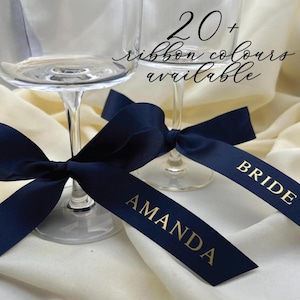 Op de afbeelding: Twee heldere champagneglazen met marineblauwe linten om de stelen gebonden. De linten hebben gouden tekst met de tekst "AMANDA" en "BRIDE". De afbeelding bevat ook de tekst "20+ lintkleuren beschikbaar".