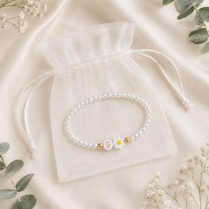 Puede incluir: Una pulsera de cuentas con pequeñas perlas blancas, una cuenta circular blanca con la letra G en dorado, una cuenta en forma de margarita blanca y amarilla, y dos pequeñas cuentas espaciadoras metálicas doradas, presentada en una bolsa blanca.