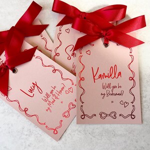 May include: Three pink cards with red ribbon bows and handwritten text. The cards say "Lucy, Will you be my Maid of Honor?", "Camilla, Will you be my Bridesmaid?" and "Sarah, Will you be my Bridesmaid?"