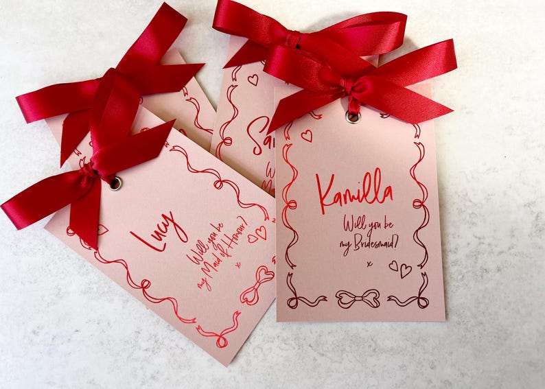 May include: Three pink cards with red ribbon bows and handwritten text. The cards say "Lucy, Will you be my Maid of Honor?", "Camilla, Will you be my Bridesmaid?" and "Sarah, Will you be my Bridesmaid?"