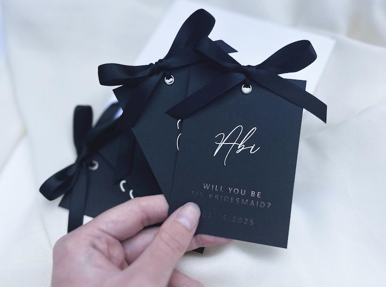 May include: Black card with white text asking "Will you be my bridesmaid?" with the name "Abi" and the date "02.12.2025" written on it. The card is tied with a black ribbon.