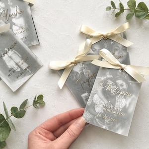 Peut inclure: Étiquettes rectangulaires en vélin translucide avec des photographies imprimées en noir et blanc, des noms en calligraphie dorée et le texte "WILL YOU BE MY BRIDESMAID?" avec une date. Chaque étiquette est surmontée d'un nœud en ruban satin crème.