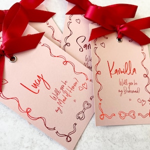 May include: Four pink cards with red ribbon bows and red ink. The cards have a decorative border and say "Lucy Will you be my Maid of Honour?", "San", "Will you be my Maid", and "Kamilla Will you be my Bridesmaid?"