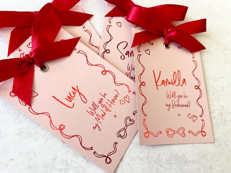 May include: Four pink cards with red ribbon bows and red ink. The cards have a decorative border and say "Lucy Will you be my Maid of Honour?", "San", "Will you be my Maid", and "Kamilla Will you be my Bridesmaid?"