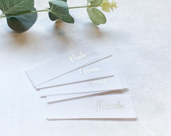 Op de afbeelding: Witte kaartjes met de woorden "Bride", "Groom", "Sarah" en "Hannah" geschreven in zilveren kalligrafie.