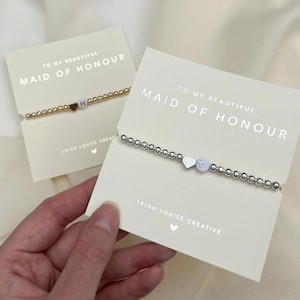 May include: Two beaded bracelets on cream-coloured display cards. One bracelet features gold-tone beads with a heart charm and the letter H. The other features silver-tone beads with a heart charm and the letter L. Both cards read TO MY BEAUTIFUL MAID OF HONOUR.