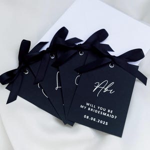 May include: Five black gift tags with white ribbon and white text. The text on the tag reads "Will you be my bridesmaid? 08.06.2025" and "Abi".