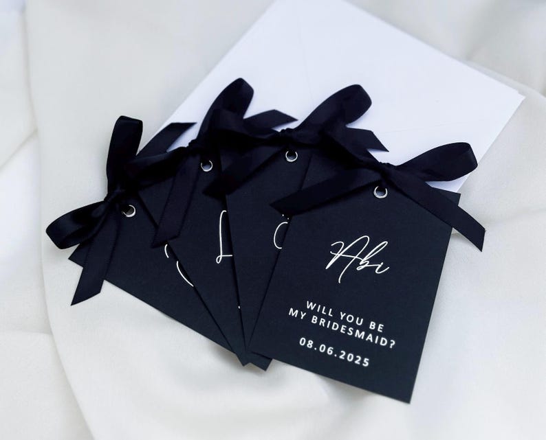 May include: Five black gift tags with white ribbon and white text. The text on the tag reads "Will you be my bridesmaid? 08.06.2025" and "Abi".