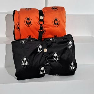 Könnte beinhalten: Zwei Hundedecken, eine orange und eine schwarze, mit einem sich wiederholenden Muster aus einer Palme und zwei Hunden. Beide Decken haben einen schwarzen Gurt mit einer Metallschnalle.