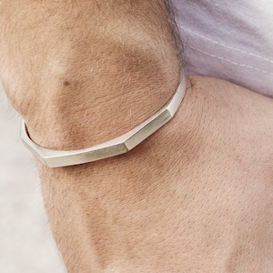 Könnte beinhalten: Ein silberfarbenes, geometrisches Armband. Das Armband hat ein facettiertes Design und wird am Handgelenk getragen. Die Person trägt ein weißes T-Shirt und eine dunkelgraue Hose. Das Bild ist eine Nahaufnahme.