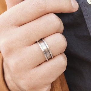 An engravable unique band ring that has its outer layers elevated in sterling silver with an oxidized finish to enhance its surface texture is shown worn by a male model.