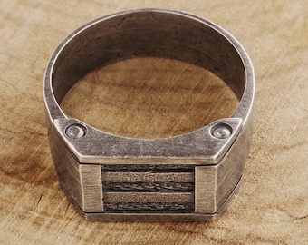 Puede incluir: Un anillo de plata con un diseño rectangular y dos pequeños remaches en los lados. El anillo está sobre una superficie de madera.