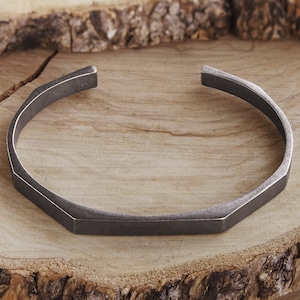 Op de afbeelding: Een zilverkleurige metalen armband met een geometrisch ontwerp. De armband is open en heeft een platte, rechthoekige vorm.