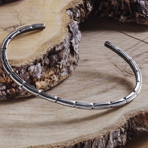 May include: A silver bangle bracelet with a textured, segmented design. The bracelet is displayed on a wooden surface.