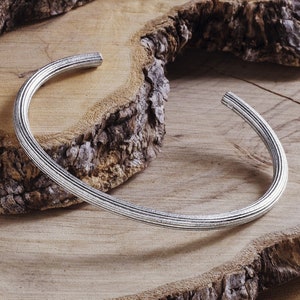 May include: A silver bangle bracelet with a textured surface. The bracelet is lying on a wooden surface.