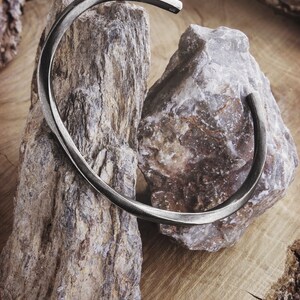 A thick, oval-shaped, square wire open bracelet with a satin finish in oxidized sterling silver is shown displayed hanging from two rocks.
