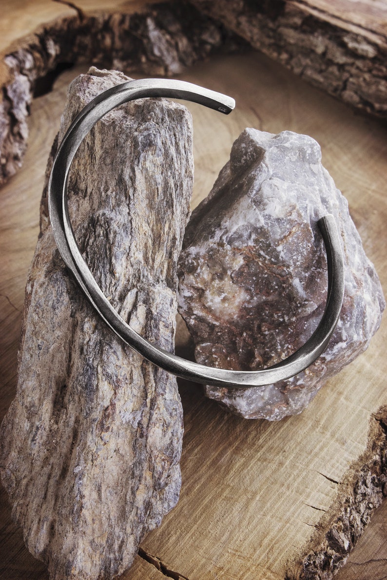 A thick, oval-shaped, square wire open bracelet with a satin finish in oxidized sterling silver is shown displayed hanging from two rocks.