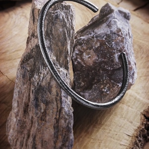 Shown hanging from two rocks, an oval-shaped open cuff bracelet made of 925 sterling silver. Its surface has a unique decorative design due to its frosted finish and oxidized to give a darker tone to the piece.