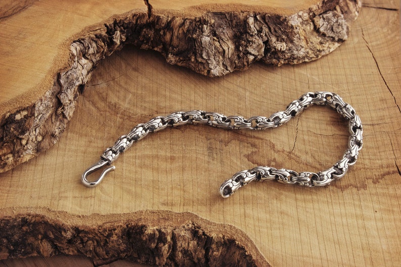 A unique chain bracelet with special beaded links with a hook clasp type closure in oxidized sterling silver that gives it a dark tone and highlights all its details is shown laid down on a wooden surface.