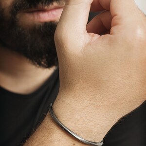 A thick, oval-shaped, square wire open bracelet with a satin finish in oxidized sterling silver is shown worn by a male model.