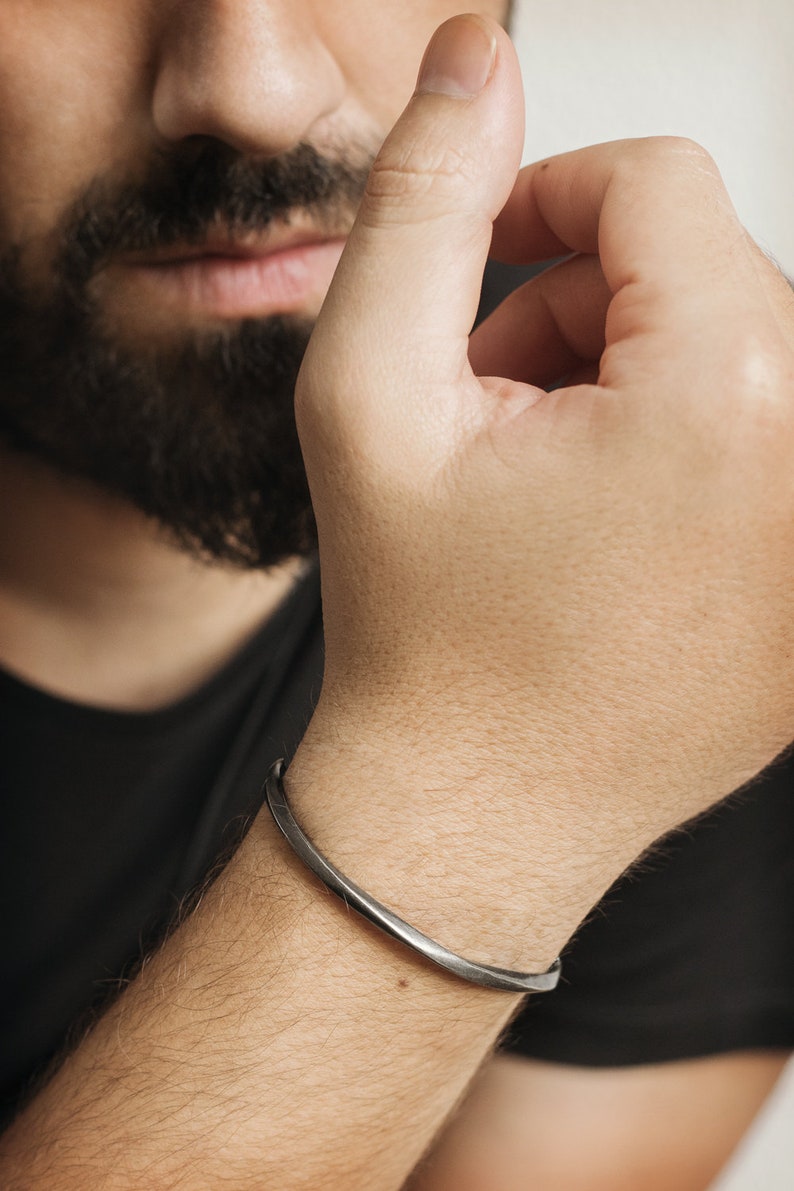 A thick, oval-shaped, square wire open bracelet with a satin finish in oxidized sterling silver is shown worn by a male model.