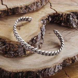 May include: A silver braided cuff bracelet on a wooden background.