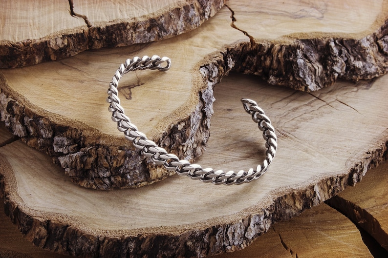 May include: A silver braided cuff bracelet on a wooden background.
