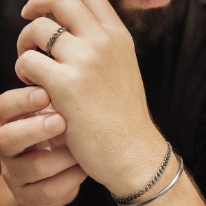 A thick, oval-shaped, square wire open bracelet with a satin finish in oxidized sterling silver is shown stacked up among other bracelets from our selection worn by a male model.