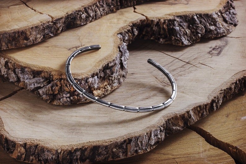 May include: A silver bangle bracelet with a textured, segmented design. The bracelet is displayed on a wooden surface.