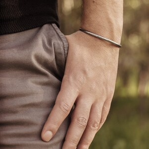 Shown worn by a male model, an oval-shaped open cuff bracelet made of 925 sterling silver. Its surface has a unique decorative design due to its frosted finish and oxidized to give a darker tone to the piece.