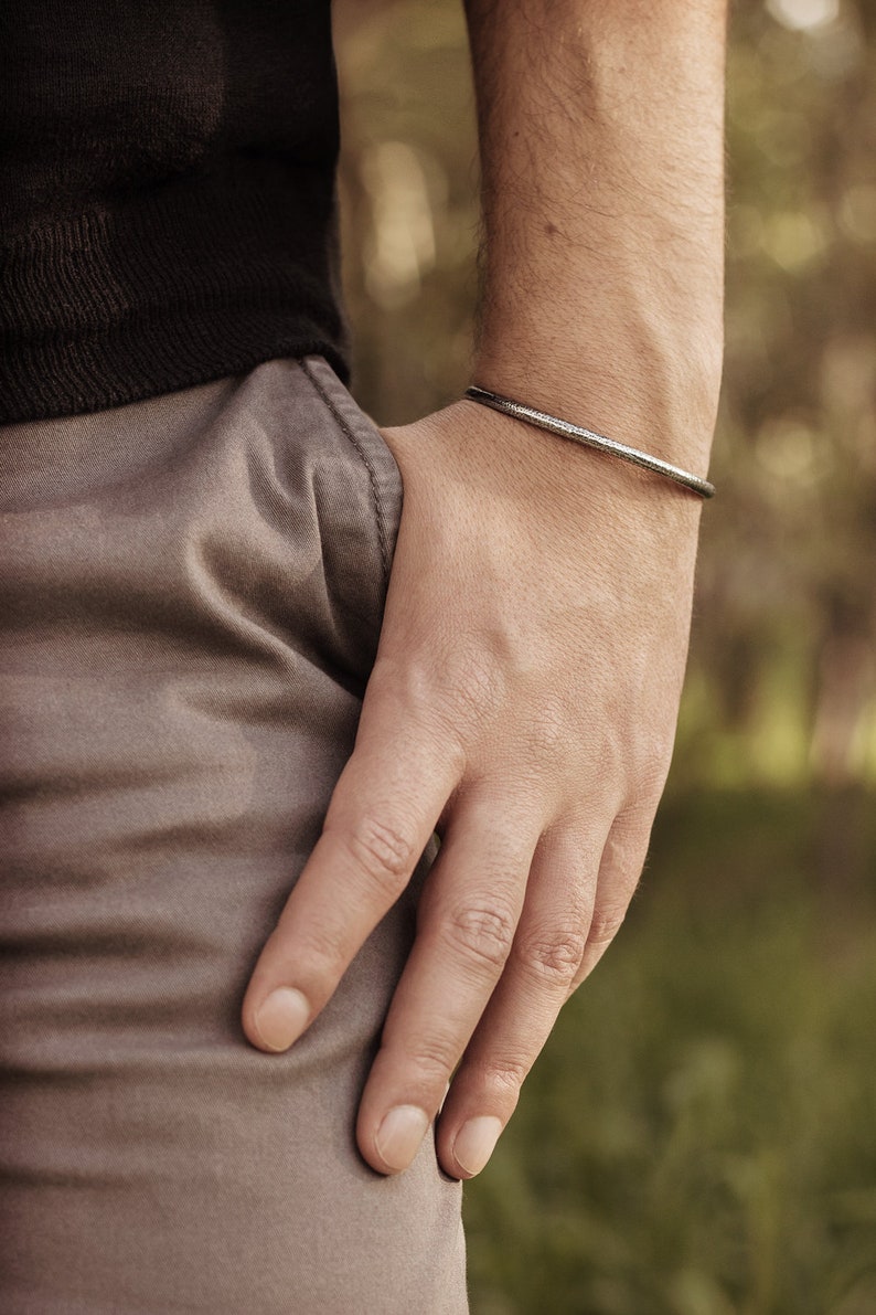 Shown worn by a male model, an oval-shaped open cuff bracelet made of 925 sterling silver. Its surface has a unique decorative design due to its frosted finish and oxidized to give a darker tone to the piece.