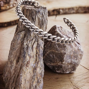 A thick, oval-shaped open cuff bracelet with a curb chain design and an oxidized finish which gives it a darker tone is shown hanging by two rocks.