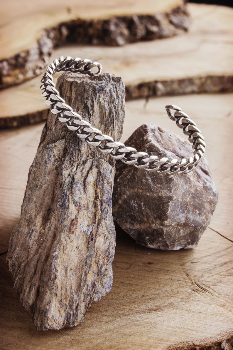 A thick, oval-shaped open cuff bracelet with a curb chain design and an oxidized finish which gives it a darker tone is shown hanging by two rocks.