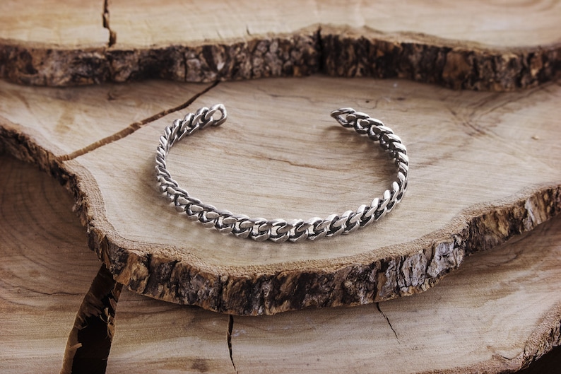 A thick, oval-shaped open cuff bracelet with a curb chain design and an oxidized finish which gives it a darker tone is shown laid down on a wooden slice.