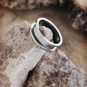 An engravable unique band ring that has its outer layers elevated in sterling silver with an oxidized finish to enhance its surface texture is shown on top of a rock.