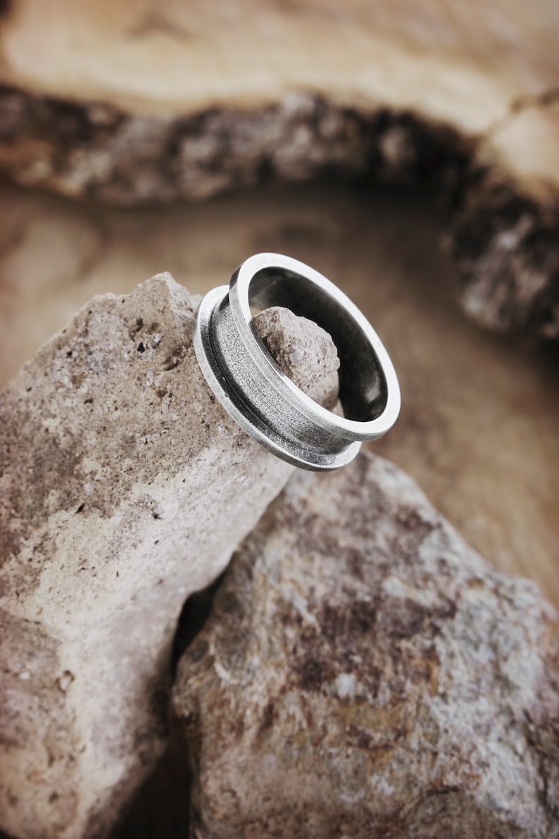 An engravable unique band ring that has its outer layers elevated in sterling silver with an oxidized finish to enhance its surface texture is shown on top of a rock.