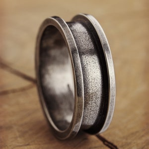 An engravable unique band ring that has its outer layers elevated in sterling silver with an oxidized finish to enhance its surface texture is shown on a wooden surface.