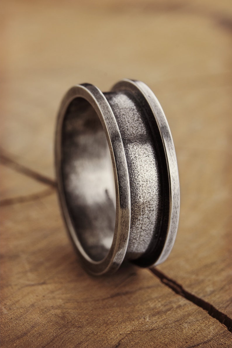 An engravable unique band ring that has its outer layers elevated in sterling silver with an oxidized finish to enhance its surface texture is shown on a wooden surface.