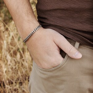 A thick, oval-shaped open cuff bracelet with a curb chain design and an oxidized finish which gives it a darker tone is shown worn by a male model.