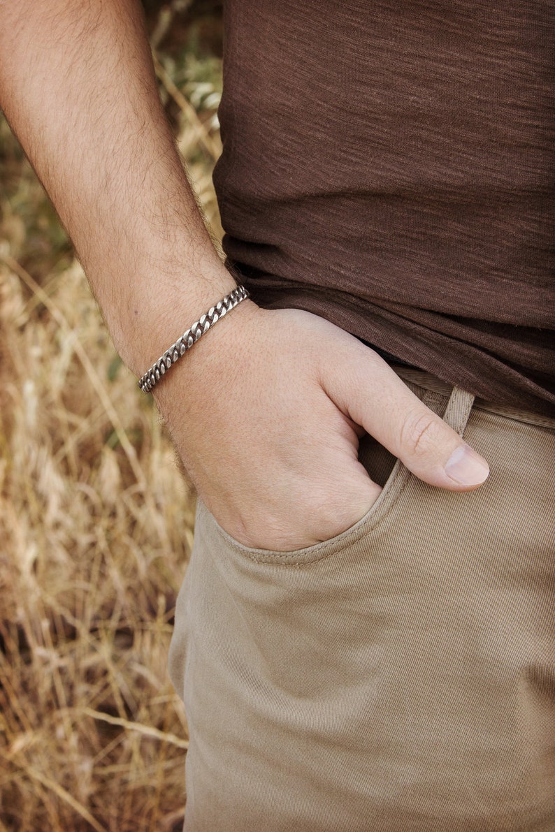 A thick, oval-shaped open cuff bracelet with a curb chain design and an oxidized finish which gives it a darker tone is shown worn by a male model.