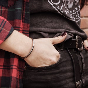 Shown worn by a female model, an oval-shaped open cuff bracelet made of 925 sterling silver. Its surface has a unique decorative design due to its frosted finish and oxidized to give a darker tone to the piece.