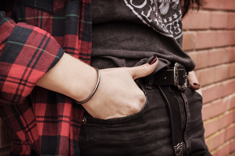 Shown worn by a female model, an oval-shaped open cuff bracelet made of 925 sterling silver. Its surface has a unique decorative design due to its frosted finish and oxidized to give a darker tone to the piece.